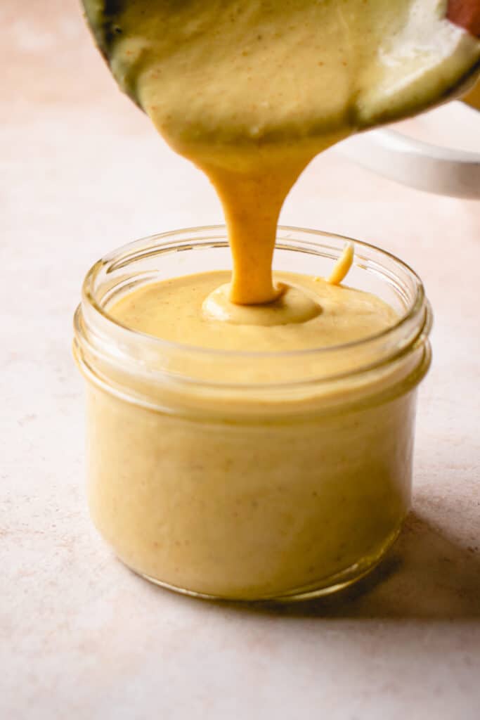 A wooden spoon pours Dairy-Free Cheese Sauce into a small glass jar.