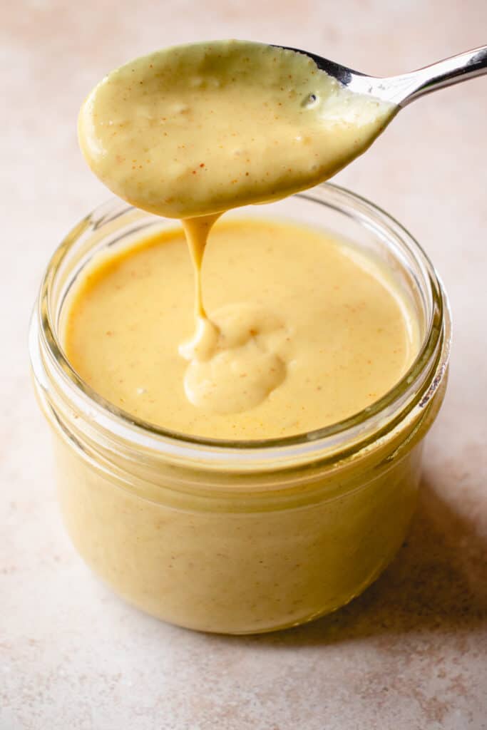 A spoon with stretchy cheese sauce dripping from it into a jar of the Dairy-Free Cheese Sauce.