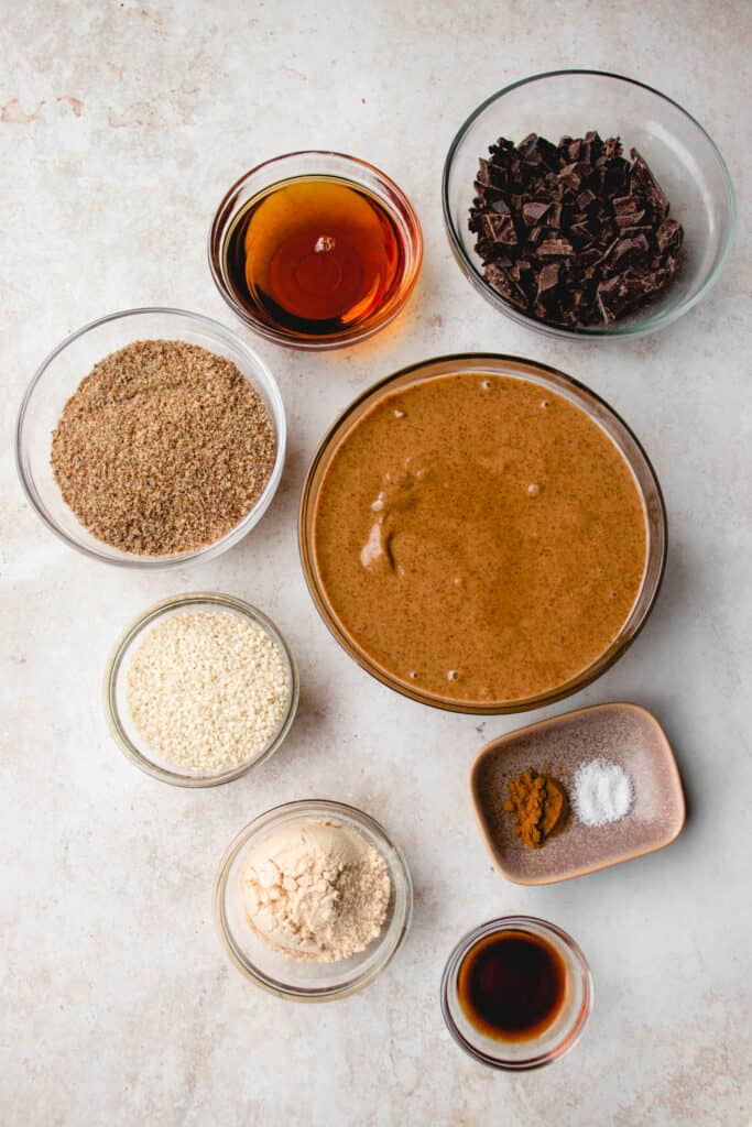 Mise en place of Protein Bar ingredients. Clockwise from top: chopped, dairy-free dark chocolate, almond butter, cinnamon and sea salt, vanilla extract, protein powder, sesame seeds, flaxseed meal, and maple syrup.