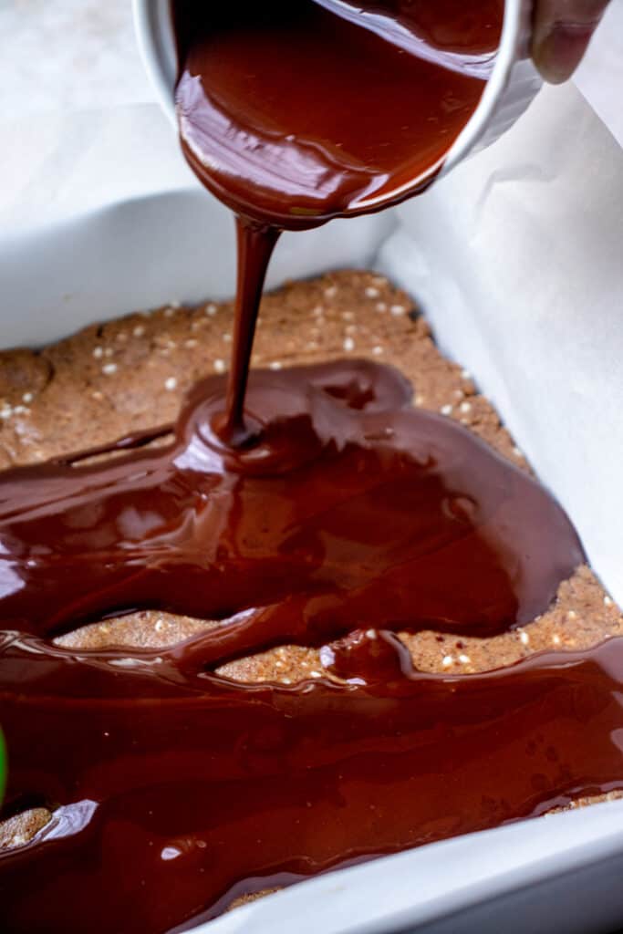 Melted chocolate is poured over the top of the protein bar mixture in a square pan lined with parchment paper.