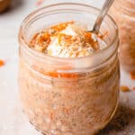 Close up of one jar with Vegan Carrot Cake Overnight Oats and a spoon. The mixture is topped with a spoonful of coconut yogurt, additional shredded carrots, sifted cinnamon, and toasted coconut flakes. The second jar is seen in the background.