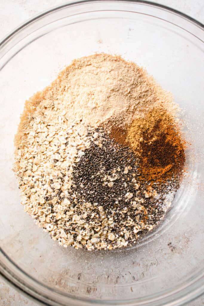A large glass bowl with gluten-free rolled oats, chia seeds, ground spices, vegan protein powder, and sea salt.