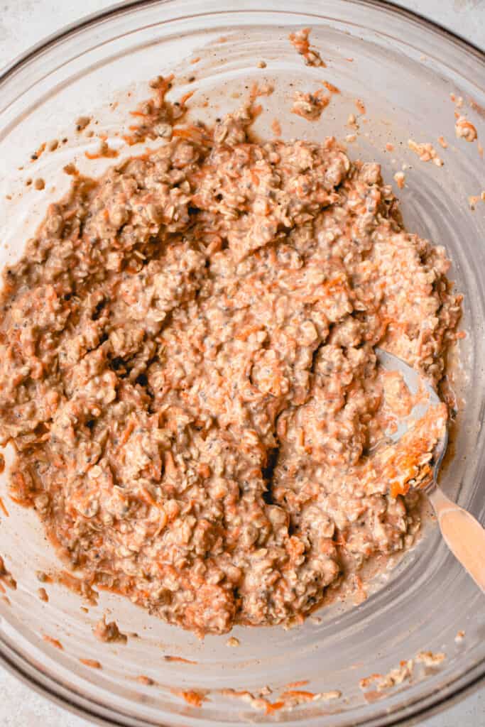 Combined shredded carrot and oat mixture, mixed with a spoon in a large glass bowl.