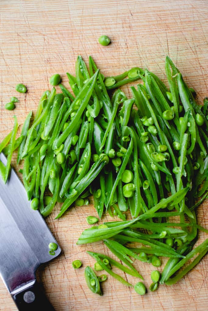 An overhead shot showing thinly sliced snap peas on a cutting board with a knife.
