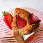 A slice of Gluten-Free Strawberry Cake with a half of a strawberry on top a red tinted dessert plate with a silver dessert fork.