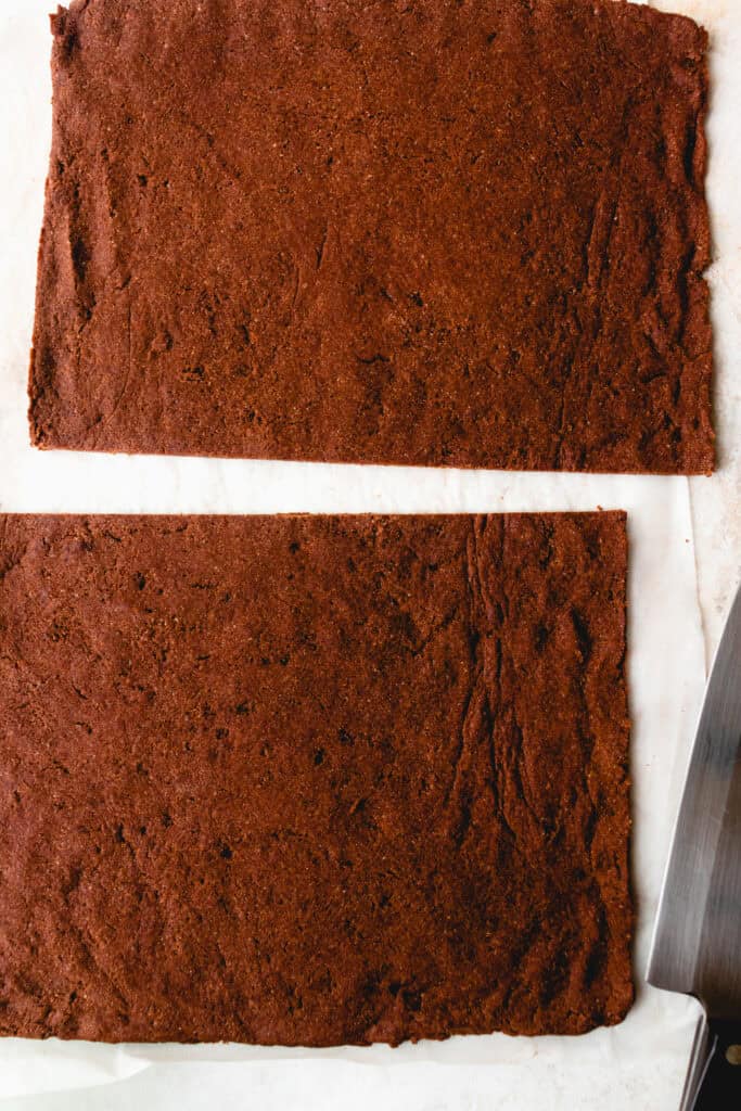 Baked chocolate sponge cake cut in half crosswise on top ph parchment paper. A sharp knife is to the bottom left of the cut pieces of cake.