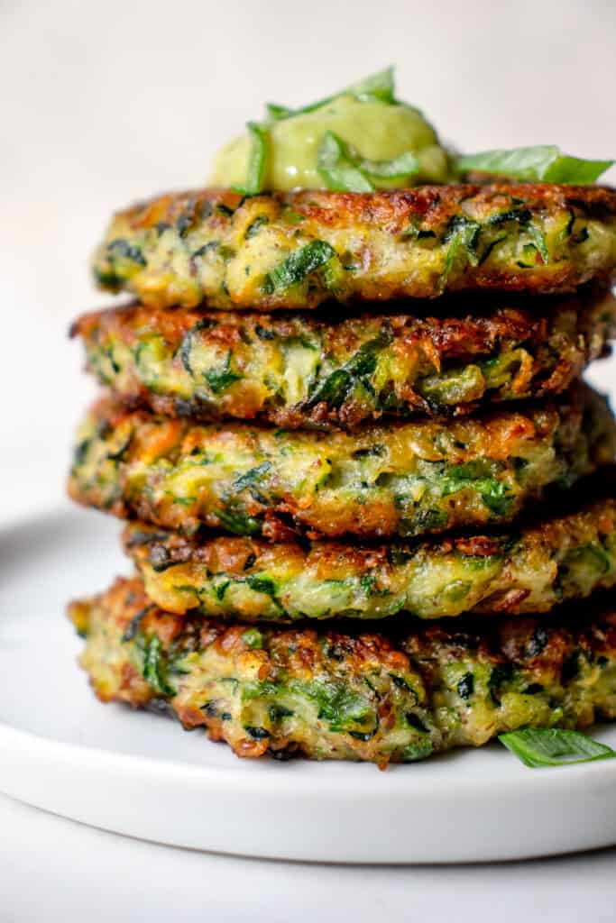 A stack of five Gluten Free Zucchini Fritters on a plate, with a dollop of Good Foods Avocado Salsa on top, garnished with green onions.