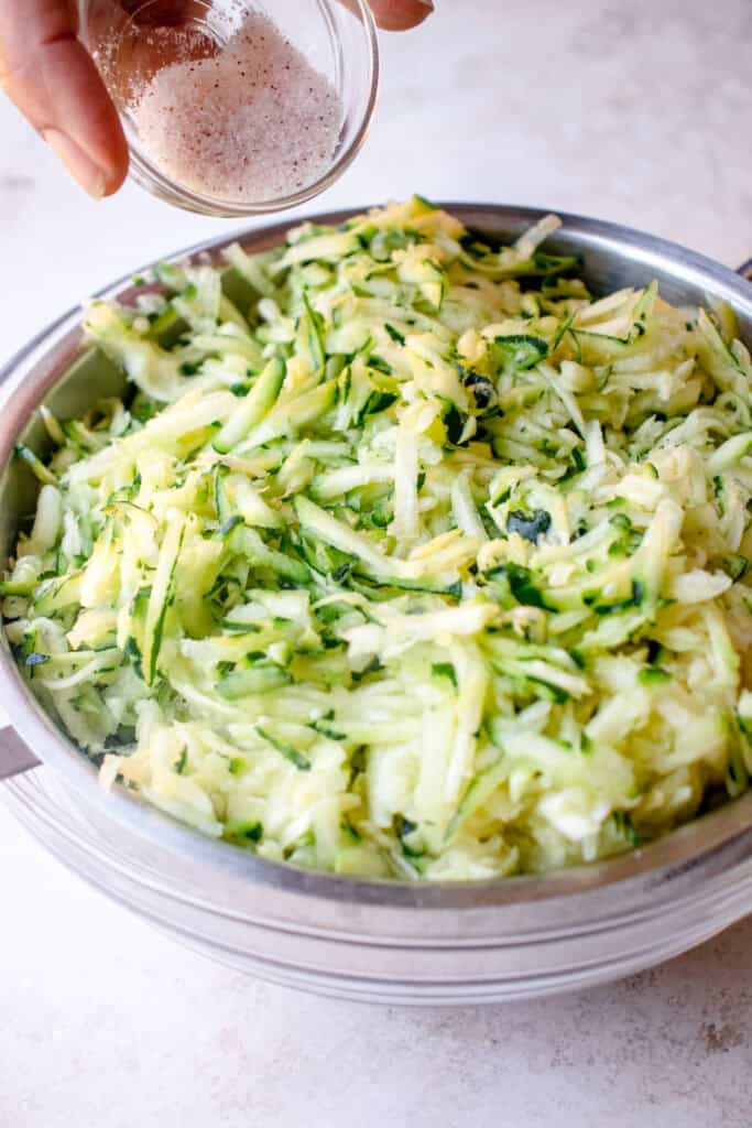 Salt being poured into shredded zucchini in a sieve, on top of a bowl.