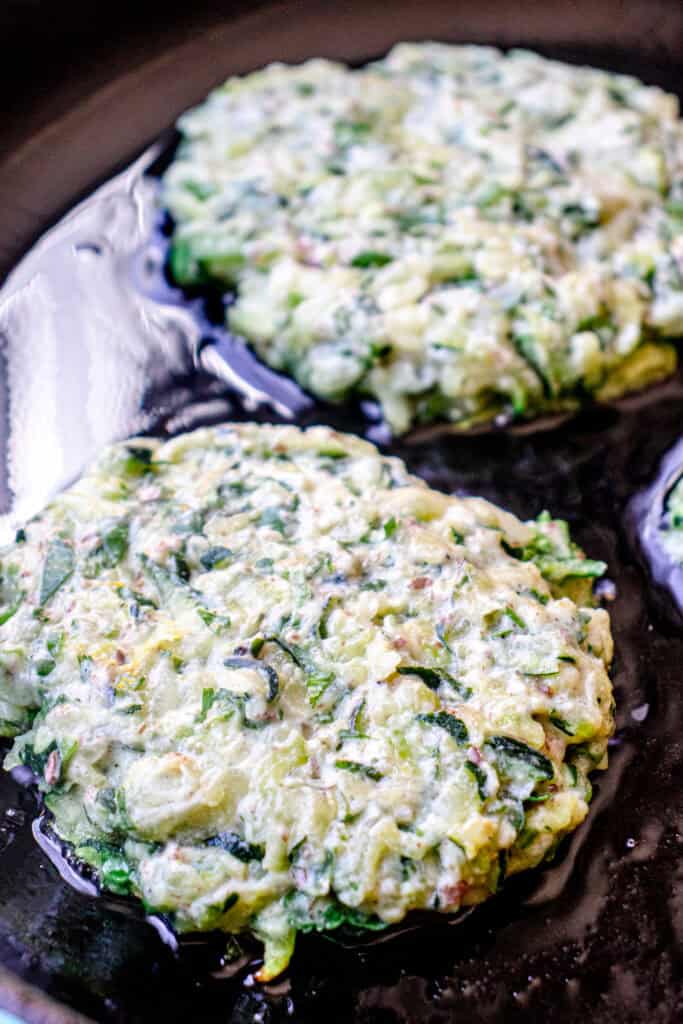 Close up of two zucchini fritter batter scoops, flattened, and frying in the skillet.