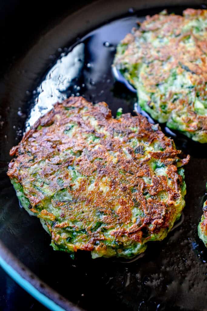 Two flipped zucchini fritters frying in a cast iron skillet, to show the cooked side is golden brown and crisp.