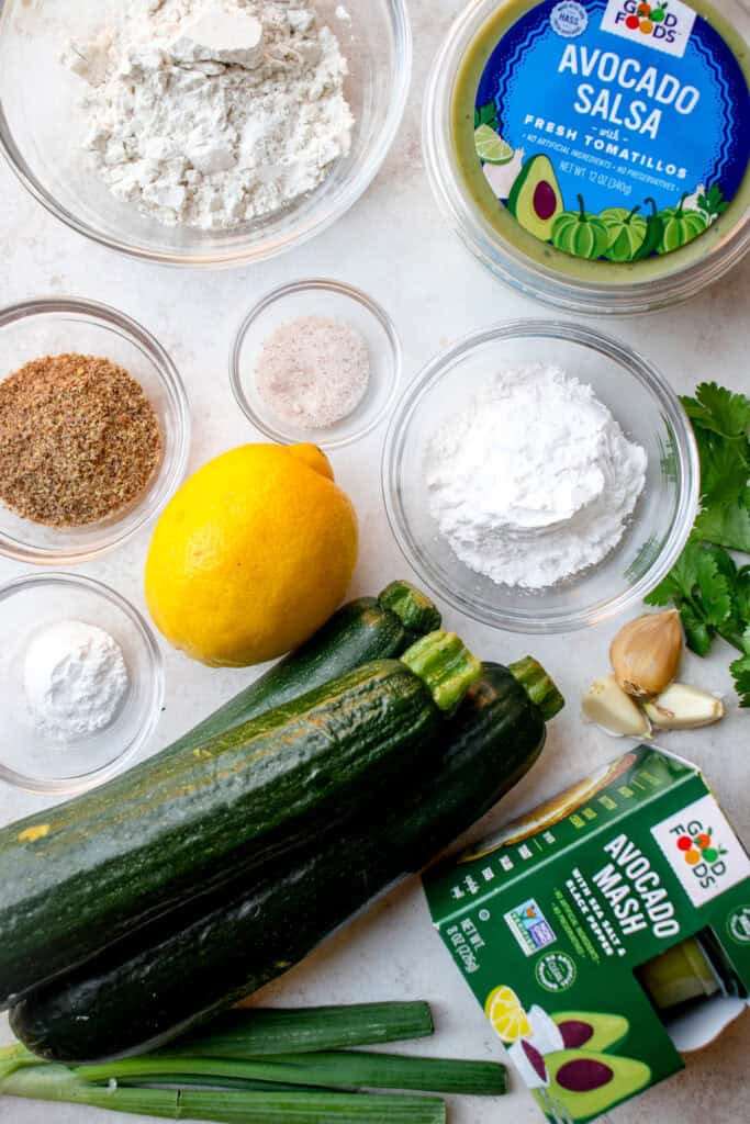 Ingredients to make Gluten Free Zucchini Fritters and the dipping sauces. Clockwise from top: Good Foods Avocado Salsa, arrowroot starch, fresh cilantro, garlic cloves, Good Foods Avocado Mash, fresh green onions, three zucchinis, baking powder, flaxseed meal, garbanzo bean flour, sea salt, and a lemon.