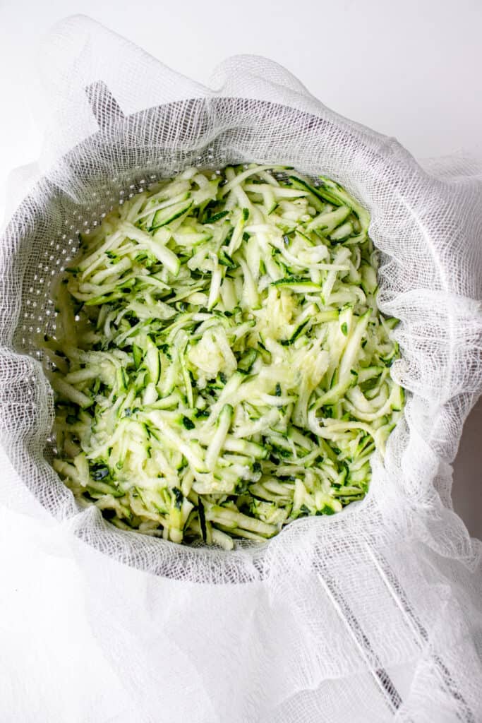 Shredded zucchini with salt in a sieve lined with a cheesecloth, on top of a bowl.