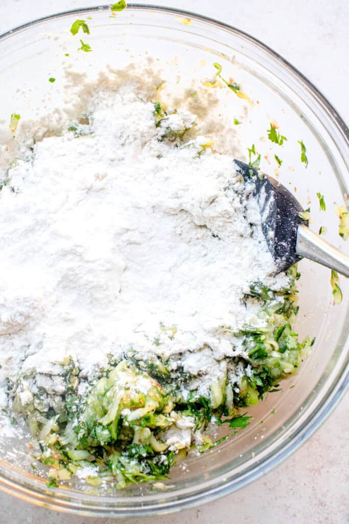 Garbanzo bean flour, arrowroot starch, and baking powder added to the seasoned zucchini in a large bowl, about to be mixed with a spatula.