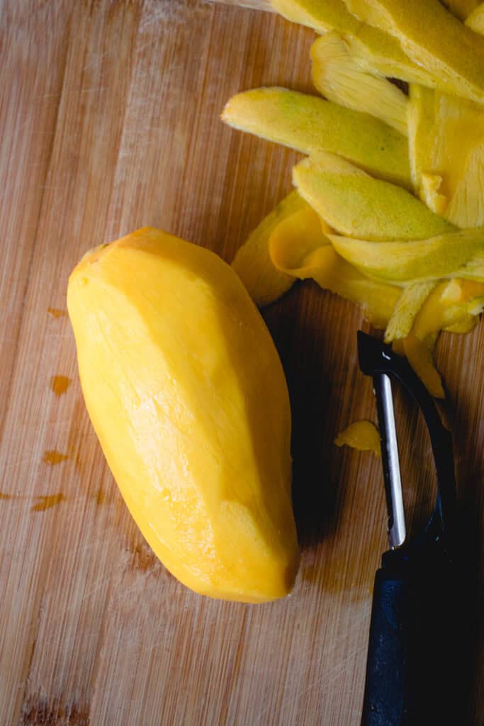 A peeled mango and a peeler on a cutting board.