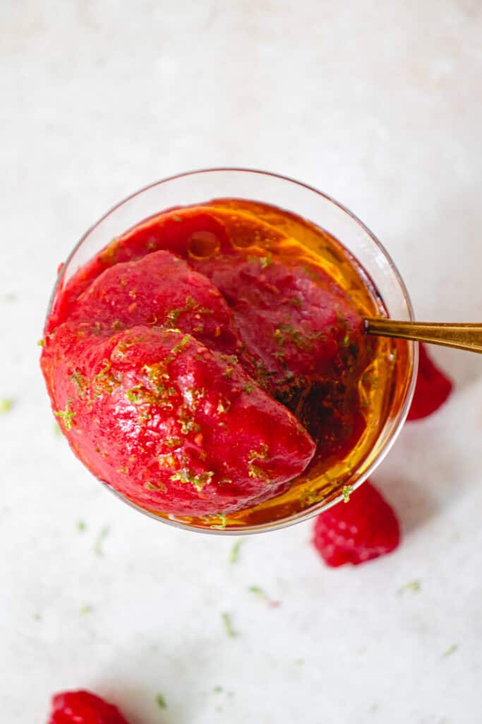 Overhead shot of a glass with Raspberry Sorbet with Oranges drizzled with olive oil and garnished with lime zest, and a gold spoon.