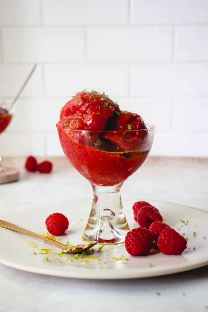 A glass with No Churn Raspberry Sorbet with Oranges, drizzled with olive oil and garnished with lime zest. The glass is on a plate with a gold spoon, with some fresh raspberries, lime zest and olive oil drippings on the plate.