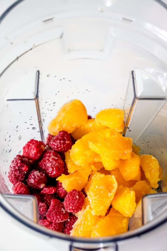 A blender bowl with frozen raspberries, frozen oranges, lemon juice, and maple syrup.