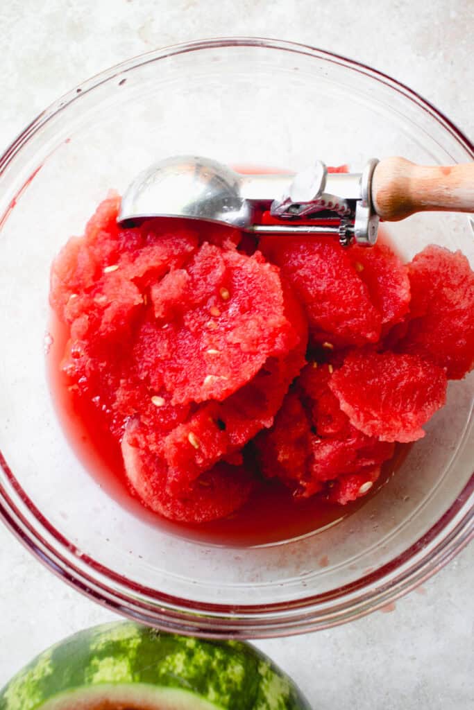 A glass bowl with watermelon flesh and an ice cream scoop.