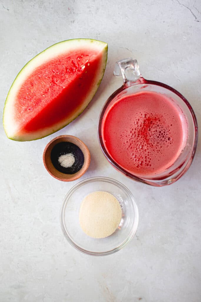 Mise en scene of Watermelon Jelly ingredients. Clockwise from top: fresh homemade watermelon juice in a measuring cup, a small bowl with gelatin, a prep bowl with some salt, and a watermelon wedge.