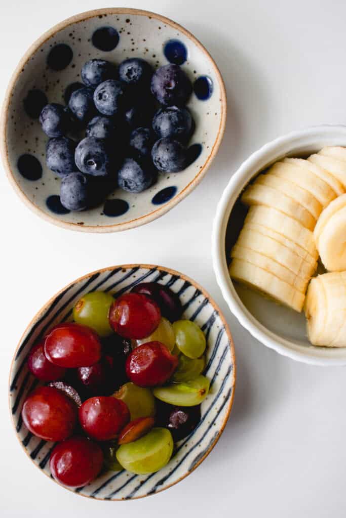 Three prep bowls with fresh fruit. Clockwise from top: sliced bananas, halved red and green grapes, and fresh blueberries.