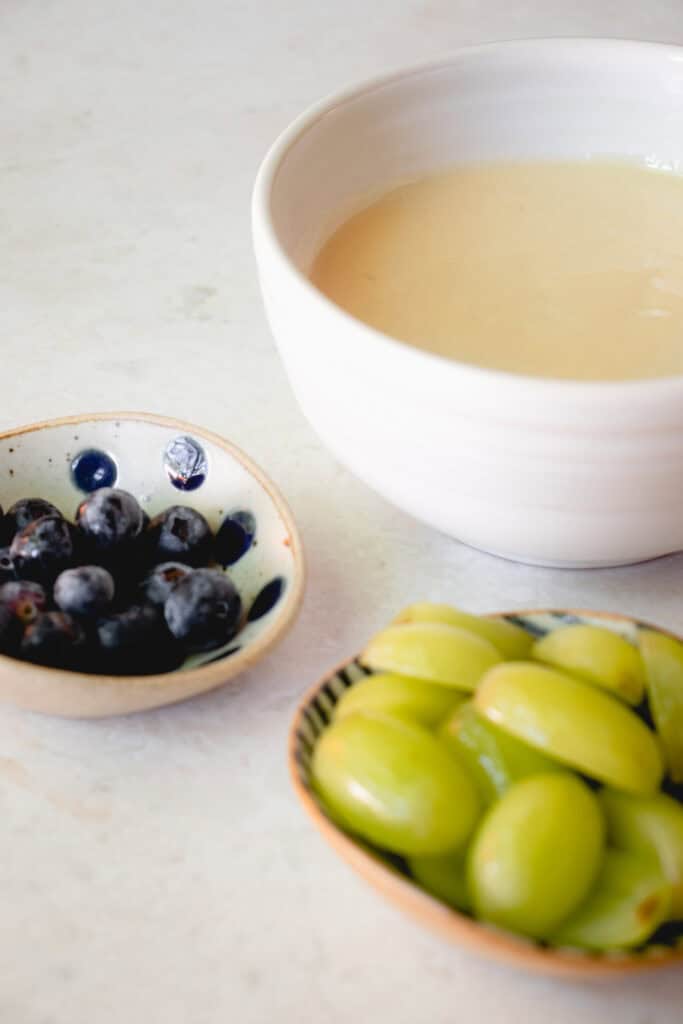 A bowl of custard, a prep dish with halved green grapes and another dish with fresh blueberries.