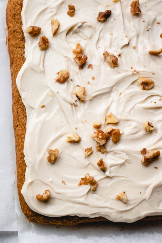 Frosted carrot cake topped with walnuts, on top of parchment paper.