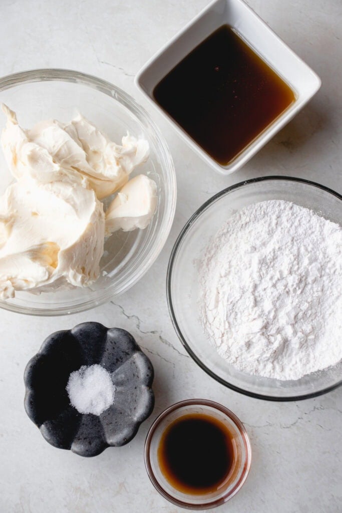 Measured frosting ingredients in bowls: maple syrup, arrowroot starch, vanilla extract, salt, and dairy-free cream cheese.