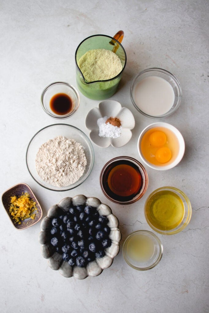 Measured out muffin ingredients in bowls: almond flour, almond milk, eggs, avocado oil, lemon juice, fresh blueberries, lemon zest, oat flour, vanilla extract, a bowl with baking powder, sea salt and cinnamon, and maple syrup.
