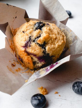 Close up of a lemon blueberry muffin with the liner unfolded, with some crumbs and blueberries scattered around it.