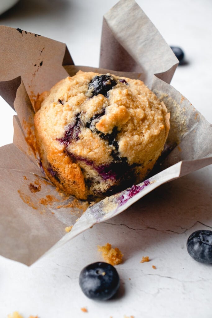 Close up of a lemon blueberry muffin with the liner unfolded, with some crumbs and blueberries scattered around it.