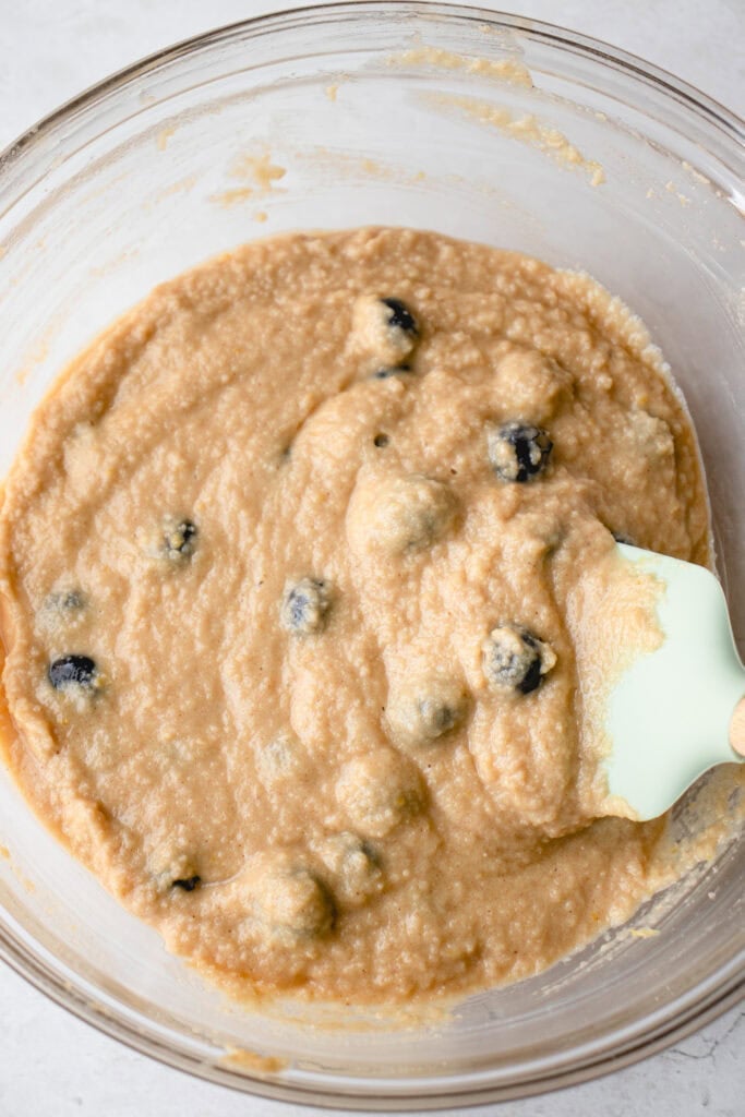 A large glass bowl with blueberries folded into muffin batter, and a spatula.