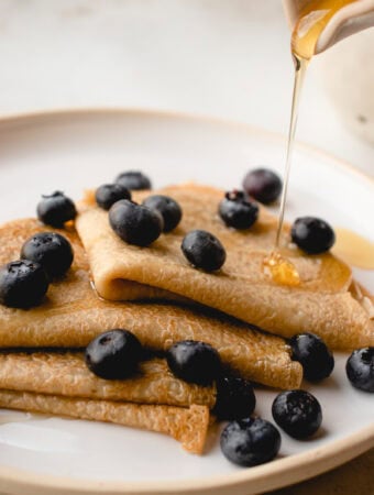 Maple syrup pours onto crepes folded in quarters with blueberries scattered on top, on a plate.